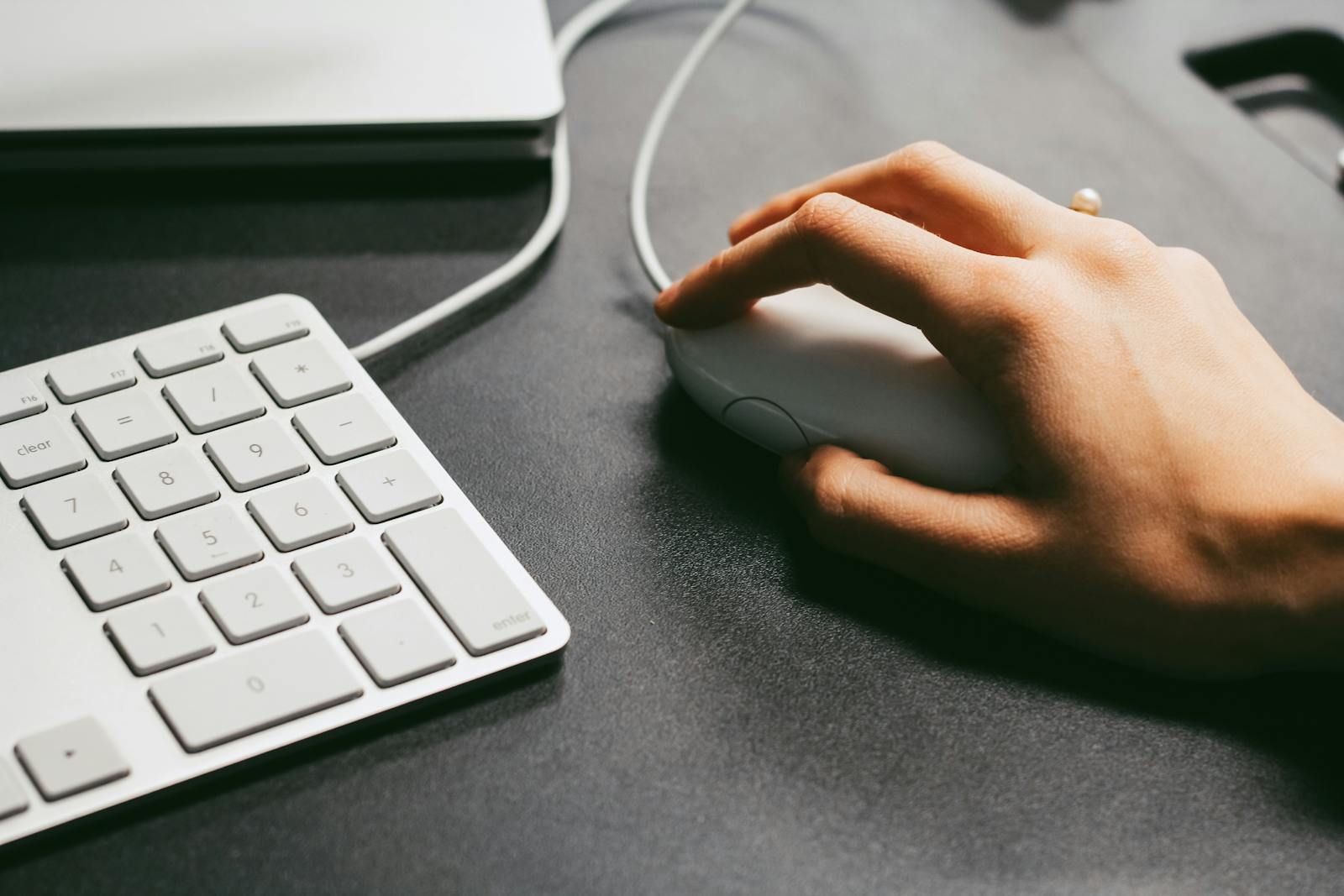Photo by Maria Stewart Hand operates a computer mouse beside a white keyboard on a dark desk, showcasing modern technology use.